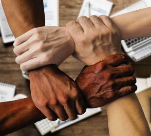 This is a photo of four arms of varying skin tones holding onto each other. Each hand is clutching a wrist, creating a perfect square, or an endless connection.