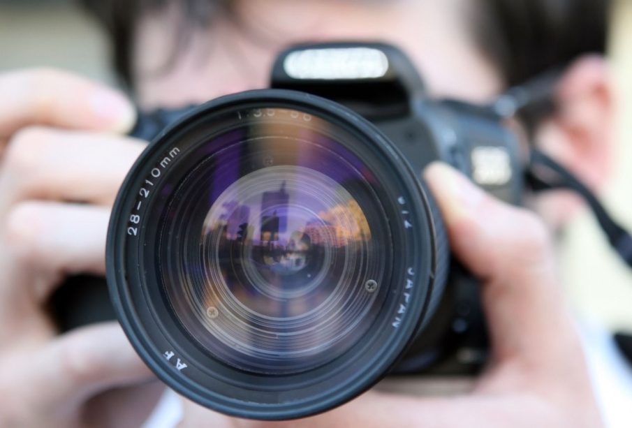 This is a close-up photo of a person holding a camera. The camera is black and the lens is front and center. A person holds the camera to their face, but the person is blurred out. 