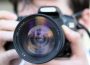 This is a close-up photo of a person holding a camera. The camera is black and the lens is front and center. A person holds the camera to their face, but the person is blurred out. 