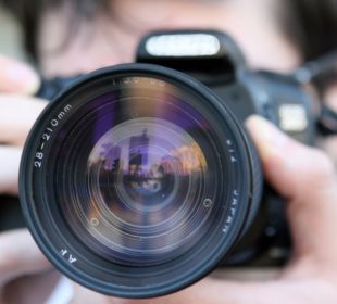 This is a close-up photo of a person holding a camera. The camera is black and the lens is front and center. A person holds the camera to their face, but the person is blurred out. 