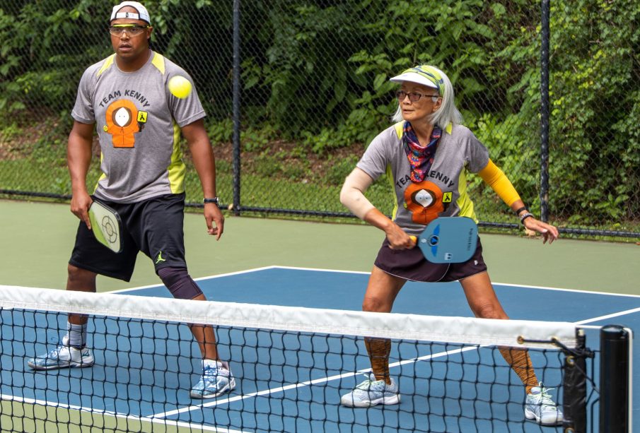 Two people stand on the same side of a pickleball court, with the one on the right having just returned the ball to the other side.