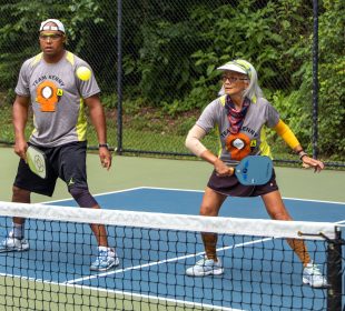 Two people stand on the same side of a pickleball court, with the one on the right having just returned the ball to the other side.
