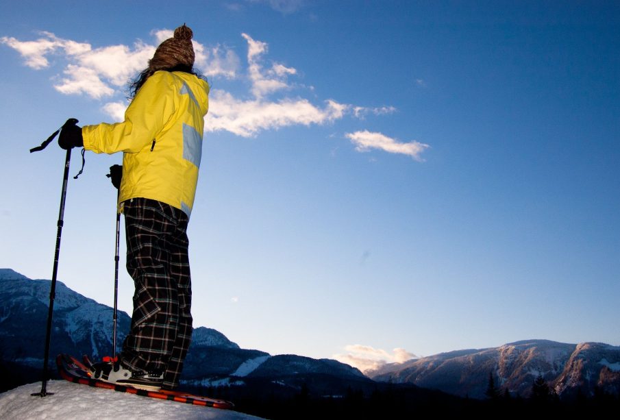 A person stands on a mountain top in their snowshoes, taking in the scenic view.