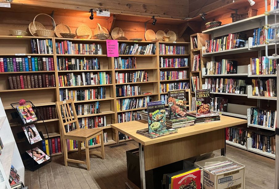 This photo shows a wooden room with wall-to-wall shelves of books. In the center is a table with a few featured cookbooks, and a few boxes of vinyl records underneath. There are also a variety of weaved baskets on top of the shelves. 