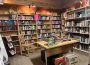 This photo shows a wooden room with wall-to-wall shelves of books. In the center is a table with a few featured cookbooks, and a few boxes of vinyl records underneath. There are also a variety of weaved baskets on top of the shelves. 