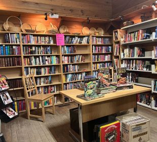 This photo shows a wooden room with wall-to-wall shelves of books. In the center is a table with a few featured cookbooks, and a few boxes of vinyl records underneath. There are also a variety of weaved baskets on top of the shelves. 