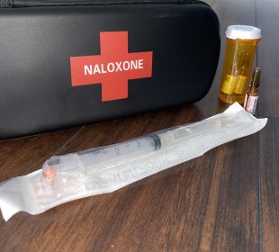 A black naloxone kit sits on a wooden table, with a still-packaged needle and three vials of the liquid injection sit beside it.
