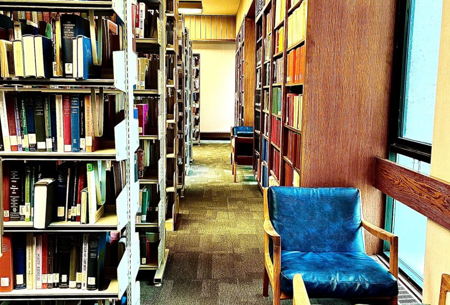 A photo looking down an aisle in Campion Library, showing shelves of books along each side and chairs beside windows to the right.