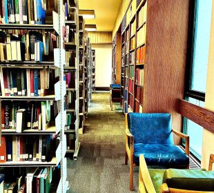 A photo looking down an aisle in Campion Library, showing shelves of books along each side and chairs beside windows to the right.