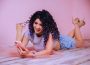 A person lays in their stomach on a bed with their feet up in the air, looking at their phone in their left hand with a smile as they play with their curly purple hair with their right.