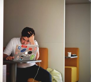 Someone sits in a booth with their head in their hand, staring at their laptop with frustration.