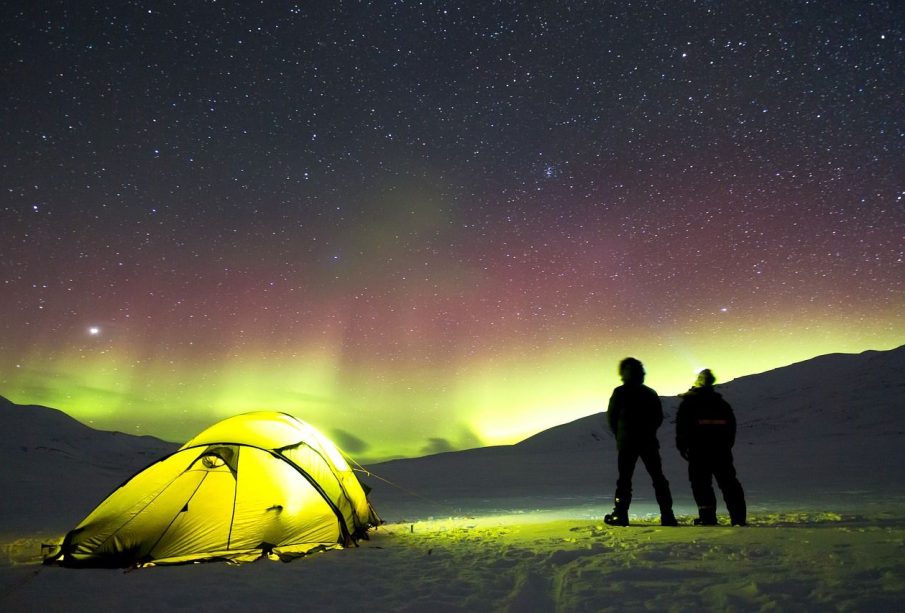 Two people stand beside their tent set up in the snow, looking at the star-filled night sky and the northern lights dancing across the horizon. 