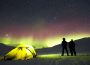 Two people stand beside their tent set up in the snow, looking at the star-filled night sky and the northern lights dancing across the horizon. 