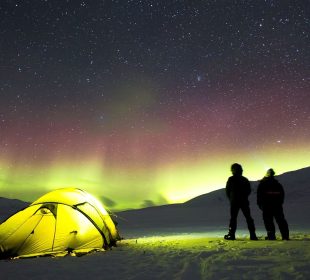 Two people stand beside their tent set up in the snow, looking at the star-filled night sky and the northern lights dancing across the horizon. 