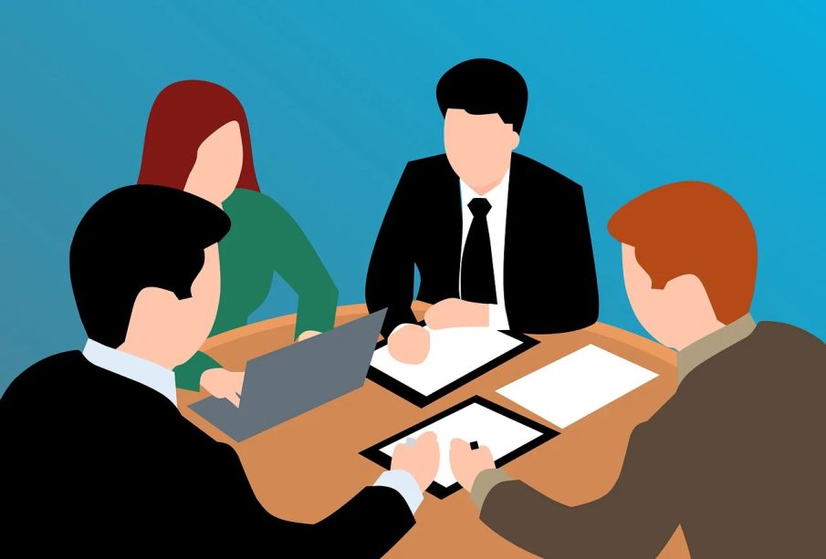 Four people sit around a table for a meeting; one person in a green shirt is typing on their laptop while the other three write their notes on paper.