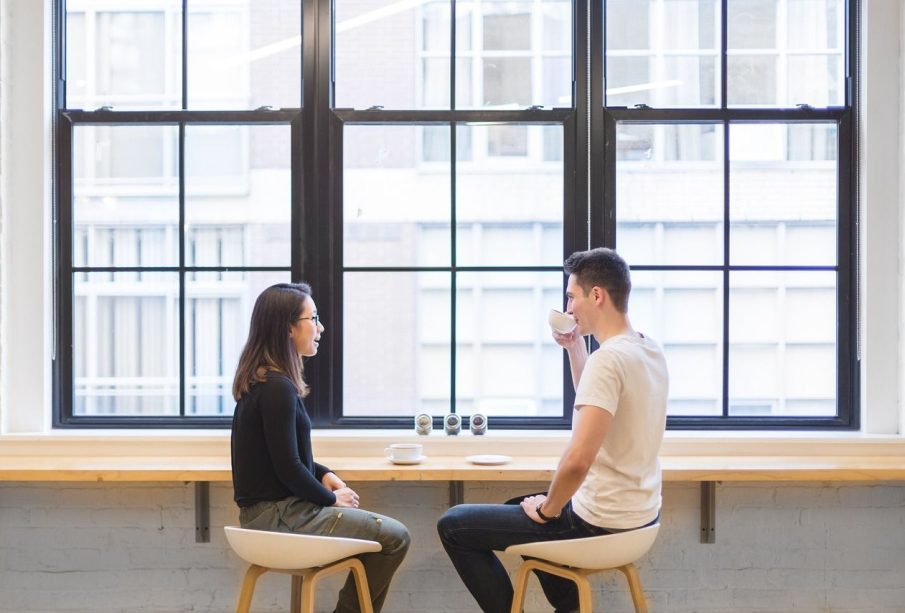 Two people sit across from each other at a cafe. One with short brown hair is sipping a drink from a mug, the other has long brown hair and sits with their hands in their lap.