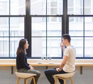 Two people sit across from each other at a cafe. One with short brown hair is sipping a drink from a mug, the other has long brown hair and sits with their hands in their lap.