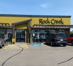 A view from the parking lot of the restaurant Rock Creek. There are three SUVs parked right by the entrance, one that’s grey on the left side with one red and one black on the right.