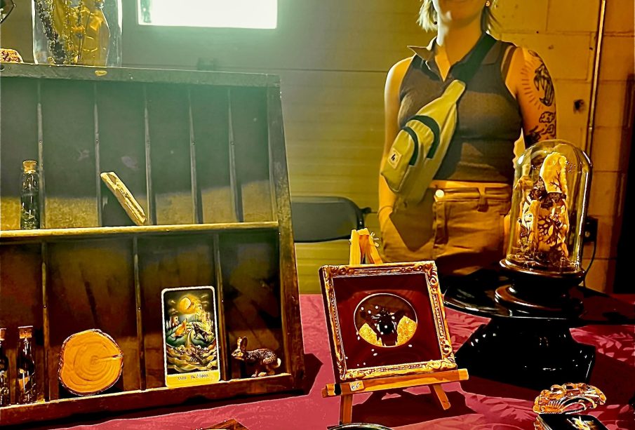 Carrie Sweeney standing to the right behind a table of her creations. There are butterflies encased in intricate wooden frames; a fox skull surrounded by orange, brown, and beige flowers typically found in a meadow encased in a glass dome; another glass dome with a butterfly on a mushroom; a lichen-covered fox skull; The Moon tarot card; and glass vials of butterfly wings with moss. There is also a magnifying glass, a small rabbit figurine, and a circle of wood also on display.