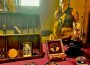 Carrie Sweeney standing to the right behind a table of her creations. There are butterflies encased in intricate wooden frames; a fox skull surrounded by orange, brown, and beige flowers typically found in a meadow encased in a glass dome; another glass dome with a butterfly on a mushroom; a lichen-covered fox skull; The Moon tarot card; and glass vials of butterfly wings with moss. There is also a magnifying glass, a small rabbit figurine, and a circle of wood also on display.