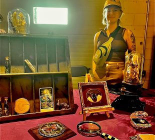 Carrie Sweeney standing to the right behind a table of her creations. There are butterflies encased in intricate wooden frames; a fox skull surrounded by orange, brown, and beige flowers typically found in a meadow encased in a glass dome; another glass dome with a butterfly on a mushroom; a lichen-covered fox skull; The Moon tarot card; and glass vials of butterfly wings with moss. There is also a magnifying glass, a small rabbit figurine, and a circle of wood also on display.