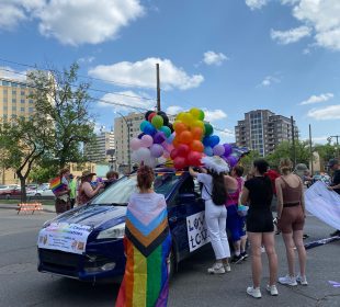 A person is wrapped in a Progressive Pride Flag (the one with a multicolor chevron-style intersection on the left of the rainbow flag) and standing near a group of people decorating a vehicle for the Pride Parade.