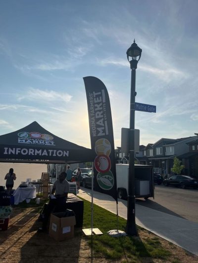 A Regina Farmers’ Market tent labelled “Information” with a cloth sign depicting “Regina Farmers’ Market” and stylistic renderings of a pod of peas, a tomato, and an ear of corn are in the mid-ground of the photo. There are boxes outside the tent and a table set up inside the tent. A person sits on a black cloth-covered table outside the tent while another in a shirt labelled “Staff” sits on a cooler to the left of the picture.