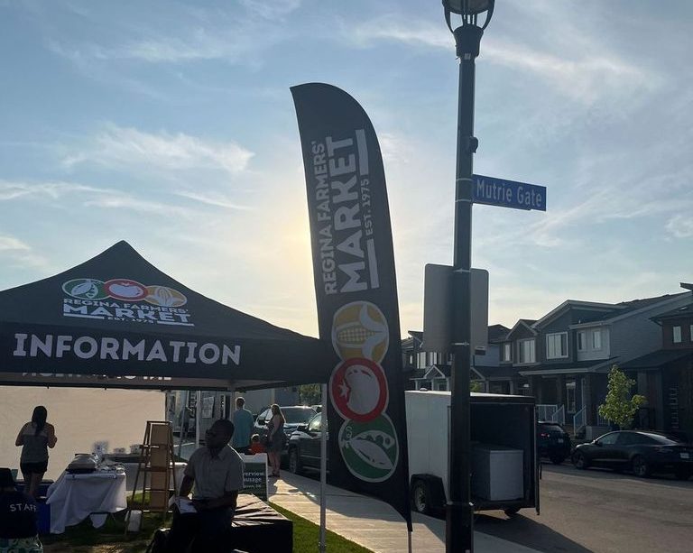 A Regina Farmers’ Market tent labelled “Information” with a cloth sign depicting “Regina Farmers’ Market” and stylistic renderings of a pod of peas, a tomato, and an ear of corn are in the mid-ground of the photo. There are boxes outside the tent and a table set up inside the tent. A person sits on a black cloth-covered table outside the tent while another in a shirt labelled “Staff” sits on a cooler to the left of the picture. 