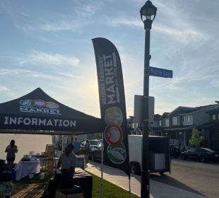 A Regina Farmers’ Market tent labelled “Information” with a cloth sign depicting “Regina Farmers’ Market” and stylistic renderings of a pod of peas, a tomato, and an ear of corn are in the mid-ground of the photo. There are boxes outside the tent and a table set up inside the tent. A person sits on a black cloth-covered table outside the tent while another in a shirt labelled “Staff” sits on a cooler to the left of the picture. 