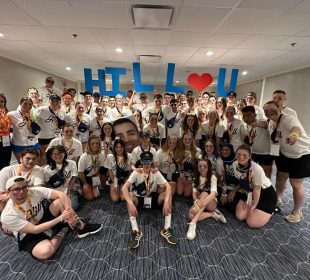 The Hill team poses for a group photo in matching team shirts and lanyards. A few members hold up signs in the back row to spell out “HILL
