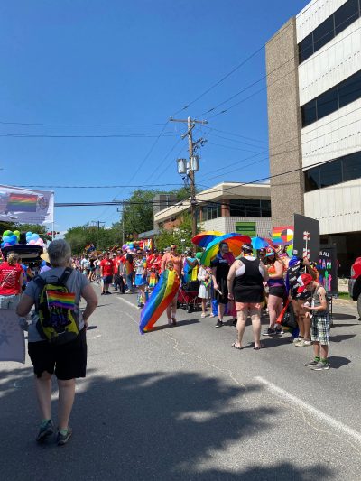 A large group of people clad in Pride flags and surrounded by many more Pride flags participating in the Queen City Pride Parade.