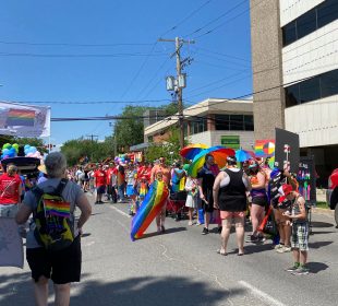 A large group of people clad in Pride flags and surrounded by many more Pride flags participating in the Queen City Pride Parade.
