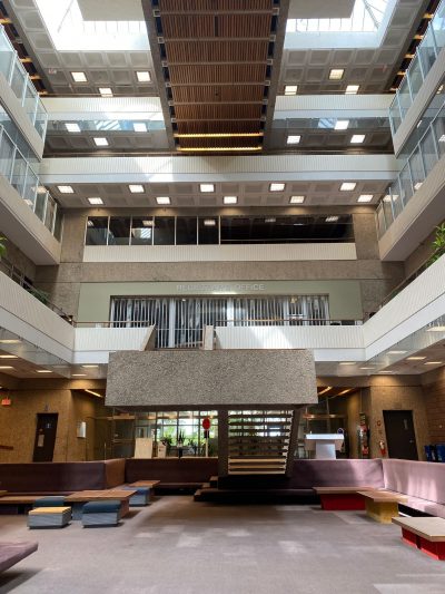 A well-lit, midday photo taken in the ‘pit’ of the Administration Humanities Building facing the main north doors, showing the full height of the building from carpeted floor to the president’s office at the top.