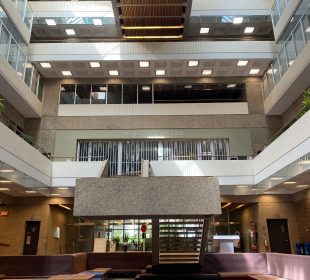 A well-lit, midday photo taken in the ‘pit’ of the Administration Humanities Building facing the main north doors, showing the full height of the building from carpeted floor to the president’s office at the top.
