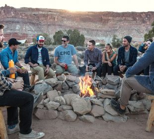 A group of 12 people sit in a valley on wooden stools around a fire surrounded by rocks to keep the fire from spreading; some people are mostly hidden by others aside from parts of their heads. There are trees, a blue van, and a canvas-topped shelter in the midground. There are mountains in the background with visible strata.