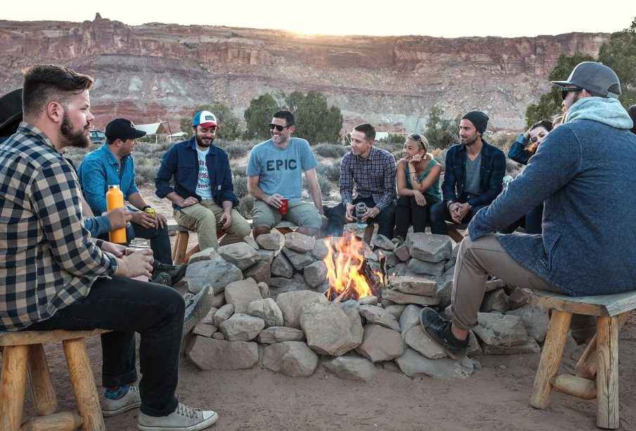 A group of 12 people sit in a valley on wooden stools around a fire surrounded by rocks to keep the fire from spreading; some people are mostly hidden by others aside from parts of their heads. There are trees, a blue van, and a canvas-topped shelter in the midground. There are mountains in the background with visible strata.