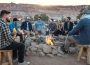 A group of 12 people sit in a valley on wooden stools around a fire surrounded by rocks to keep the fire from spreading; some people are mostly hidden by others aside from parts of their heads. There are trees, a blue van, and a canvas-topped shelter in the midground. There are mountains in the background with visible strata.