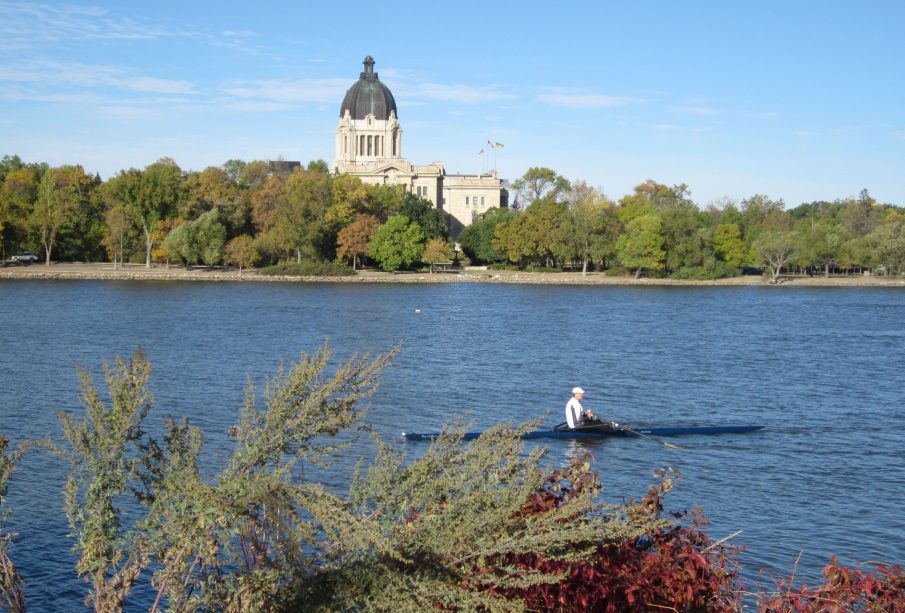 A person rowing a boat on Wascana lake on a sunny day. The Legislative Building is visible in the background.