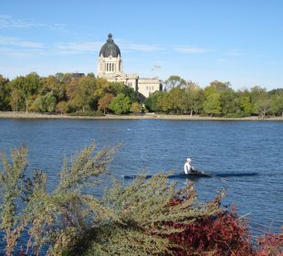 A person rowing a boat on Wascana lake on a sunny day. The Legislative Building is visible in the background.