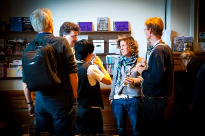 A group of five people stand in a foyer, apparently during the intermission of an event, sharing conversation and drinks before the curtain rises again.