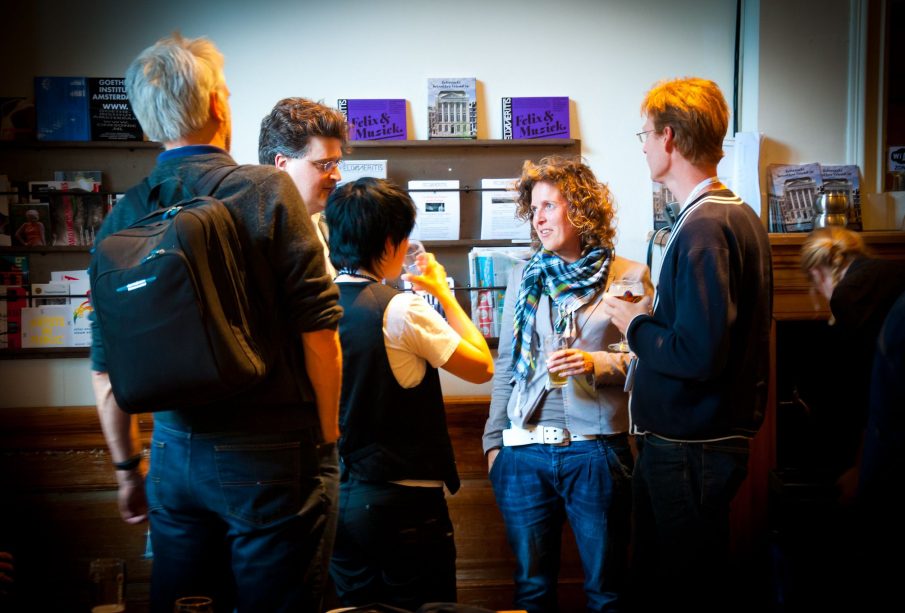 A group of five people stand in a foyer, apparently during the intermission of an event, sharing conversation and drinks before the curtain rises again.
