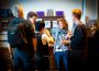 A group of five people stand in a foyer, apparently during the intermission of an event, sharing conversation and drinks before the curtain rises again.