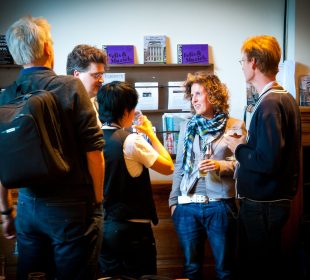 A group of five people stand in a foyer, apparently during the intermission of an event, sharing conversation and drinks before the curtain rises again.