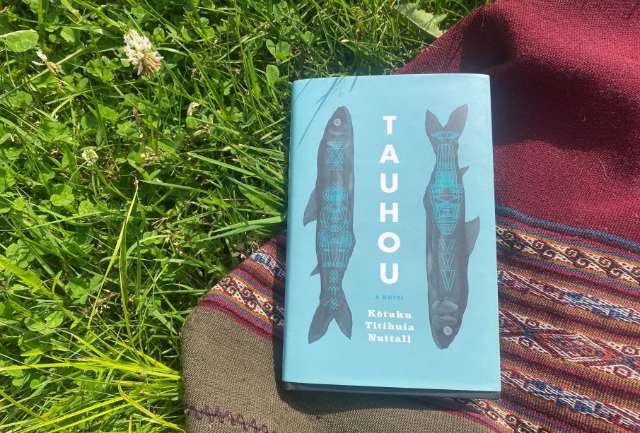 A blue book lays on top of a red woven blanket on a patch of grass with some flowers. The book’s cover is its title (Tauhou) written down the middle of the cover in bold black lettering, and the author’s name (Kōtuku Titihuia Nuttall) is written below the title in three lines, and there are two black fish with blue overlaid designs along the sides of the title.
