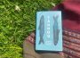 A blue book lays on top of a red woven blanket on a patch of grass with some flowers. The book’s cover is its title (Tauhou) written down the middle of the cover in bold black lettering, and the author’s name (Kōtuku Titihuia Nuttall) is written below the title in three lines, and there are two black fish with blue overlaid designs along the sides of the title.