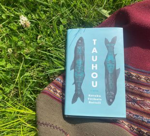 A blue book lays on top of a red woven blanket on a patch of grass with some flowers. The book’s cover is its title (Tauhou) written down the middle of the cover in bold black lettering, and the author’s name (Kōtuku Titihuia Nuttall) is written below the title in three lines, and there are two black fish with blue overlaid designs along the sides of the title.
