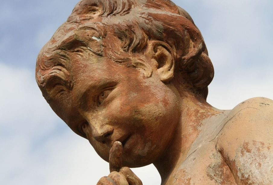 A yellowed stone statue of a child with curly hair holds its finger to its lips in a hushing motion.