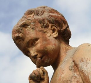 A yellowed stone statue of a child with curly hair holds its finger to its lips in a hushing motion.