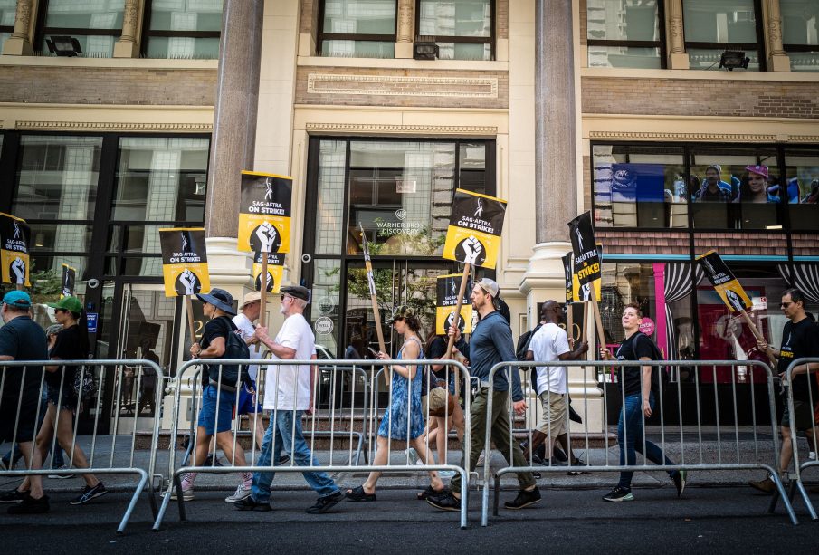 A group of striking workers holding signs stating “SAG-AFTRA ON STRIKE!” walking outside a brick building belonging to Warner Bros. Discovery. They are walking partially on the street and partially on the sidewalk, protected from cars by a mobile metal fence.