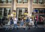 A group of striking workers holding signs stating “SAG-AFTRA ON STRIKE!” walking outside a brick building belonging to Warner Bros. Discovery. They are walking partially on the street and partially on the sidewalk, protected from cars by a mobile metal fence.